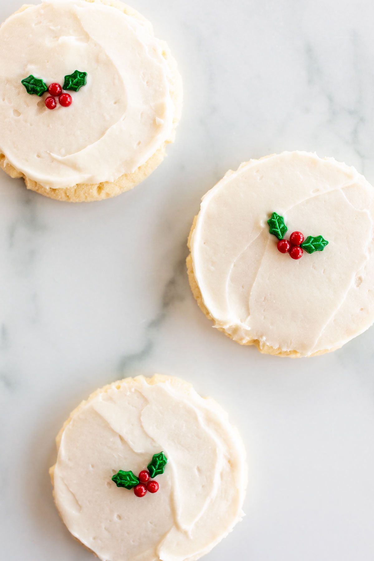 Christmas sugar cookies on a marble surface, with white frosting and a simple holly berry decoration in the center.