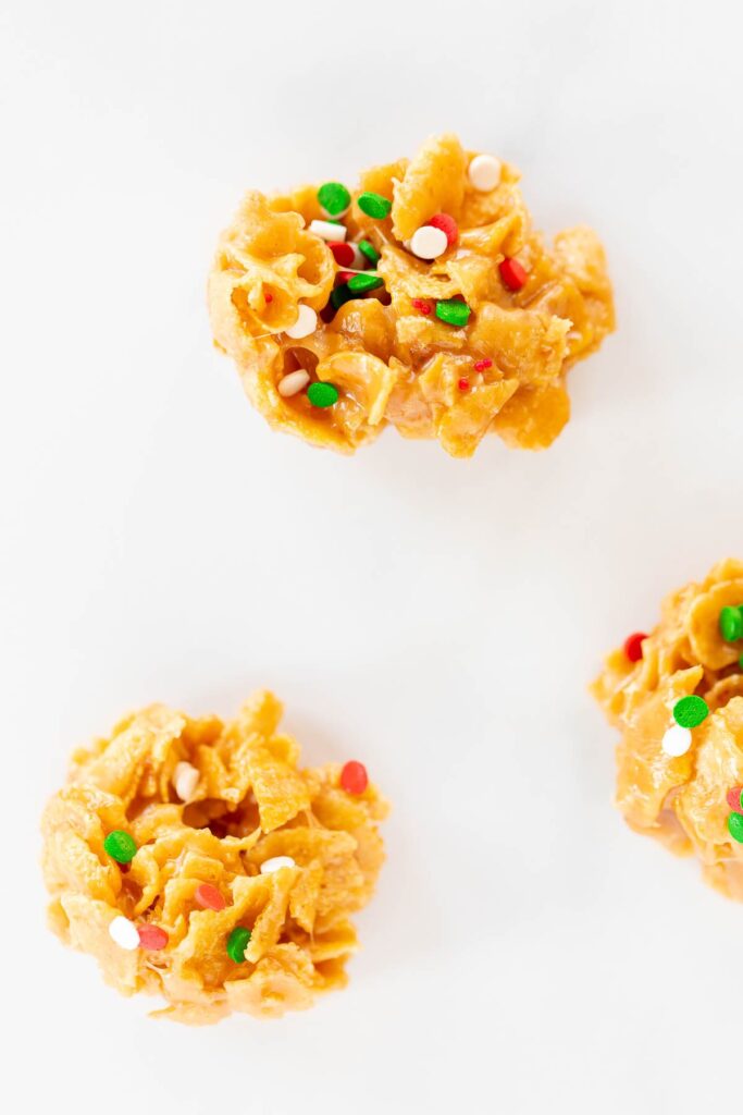Three cornflake cookies with Christmas sprinkles on a white surface.