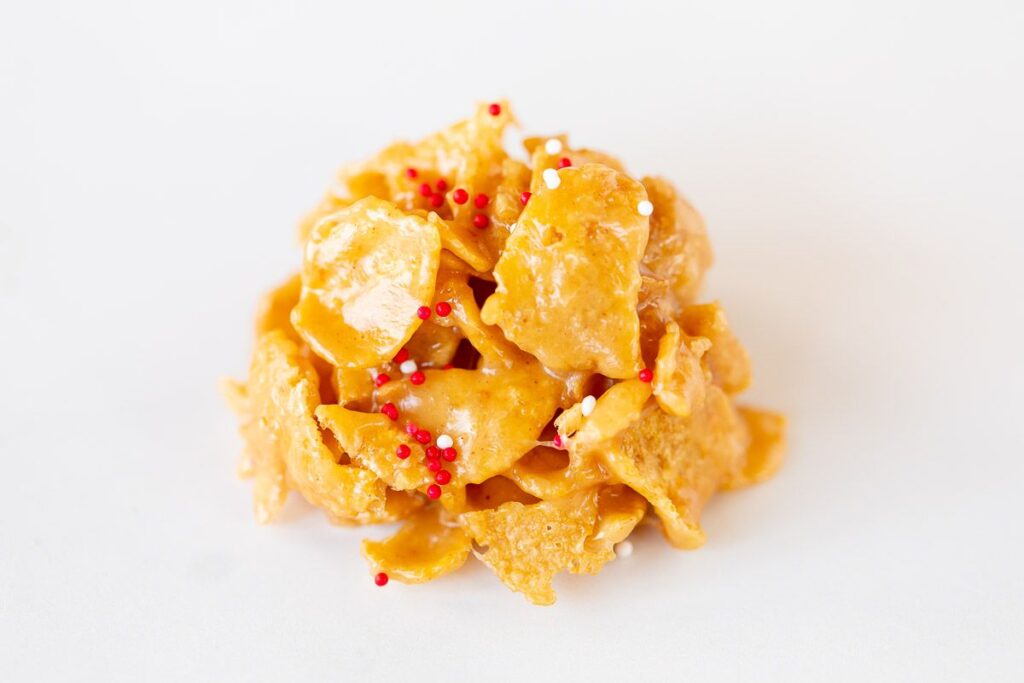 A single no bake cornflake cookie on a white surface, topped with Christmas sprinkles.