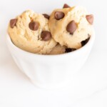 A white bowl on a white surface with scoops of an edible cookie dough recipe with chocolate chips.