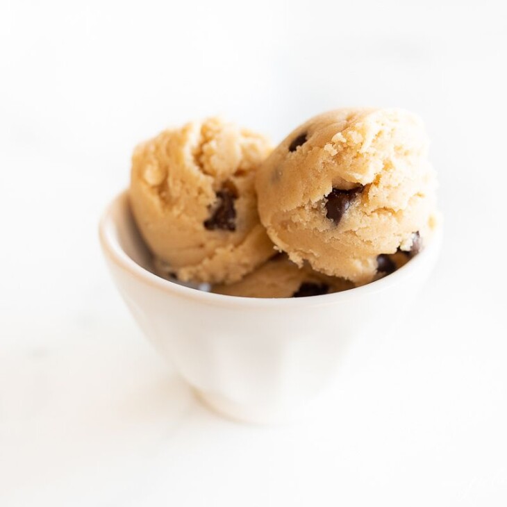 A white bowl on a white surface with balls of an edible cookie dough recipe with chocolate chips.