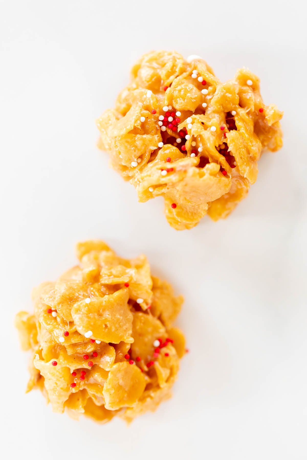 A pair of no bake cornflake cookies on a white surface, topped with Christmas sprinkles.