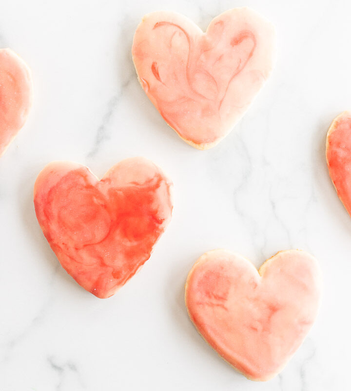 Sugar cookies in a cutout heart shape place on a marble surface, topped with pink royal icing.