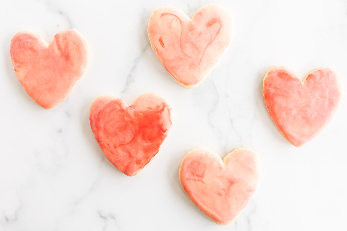 Sugar cookies in a cutout heart shape place on a marble surface, topped with pink royal icing.