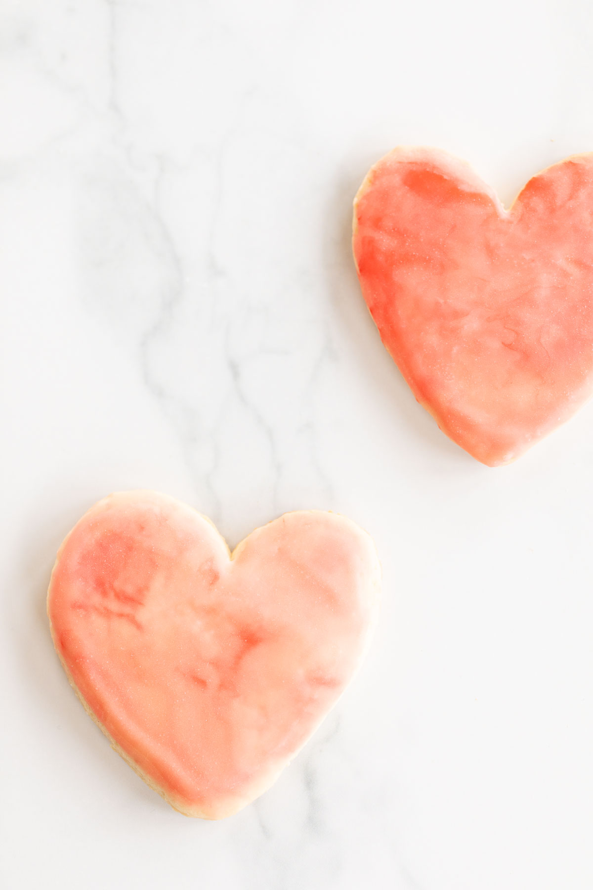 Sugar cookies in a cutout heart shape place on a marble surface, topped with pink royal icing.