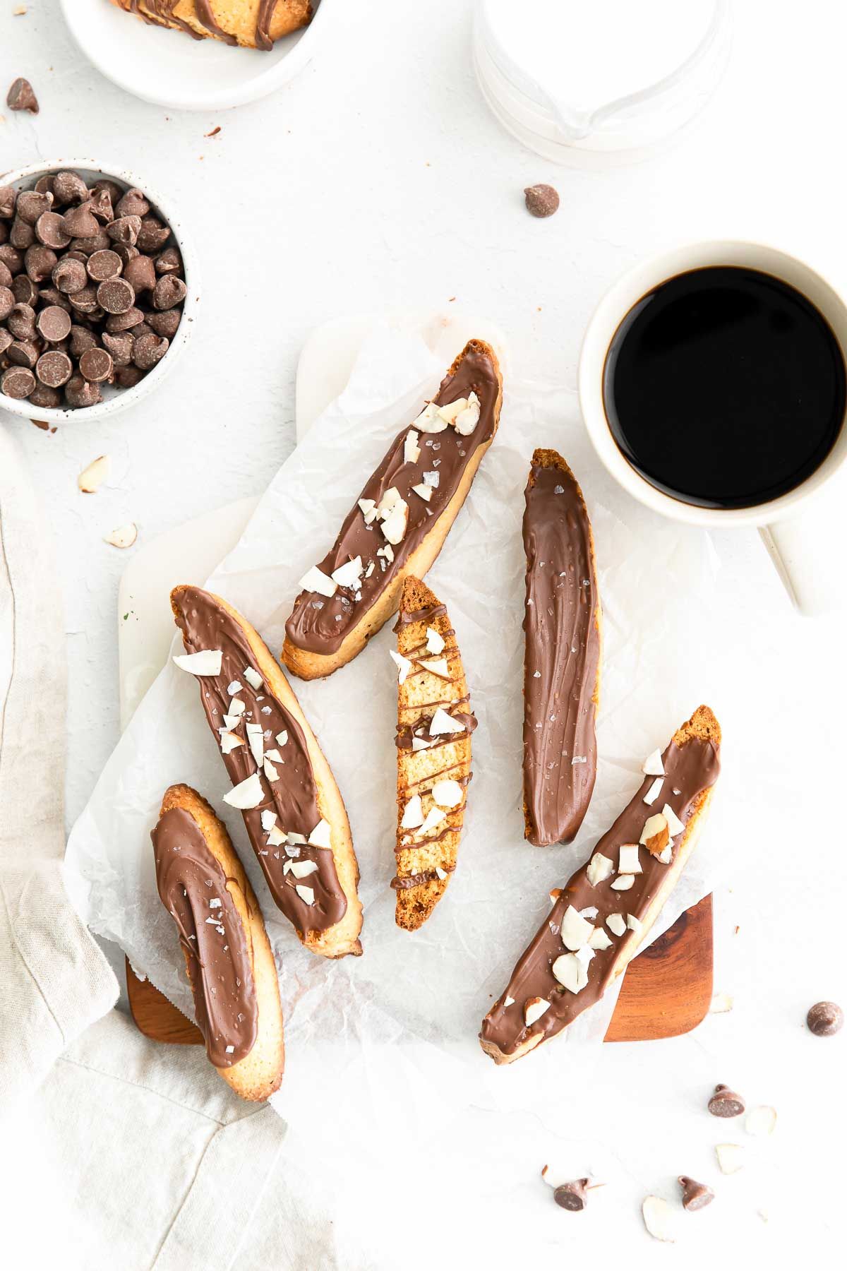 Vanilla biscotti covered in chocolate on a marble surface, bowl of chocolate chips and cup of coffee in the background.