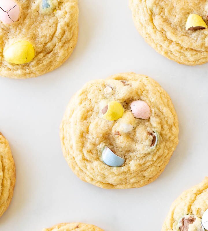 Mini egg cookies with pastel colored candy eggs spread out on a marble surface.