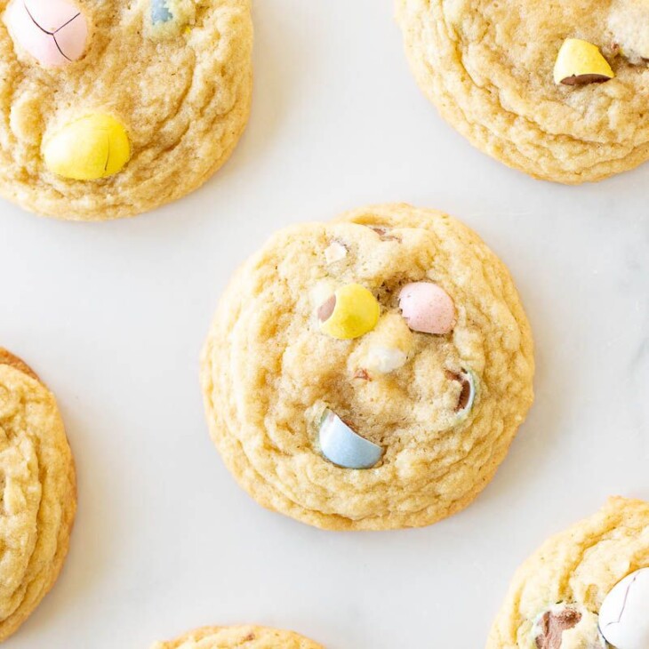 Mini egg cookies with pastel colored candy eggs spread out on a marble surface.