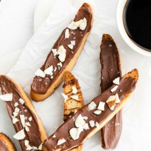 Vanilla biscotti covered in chocolate on a marble surface, bowl of chocolate chips and cup of coffee in the background.
