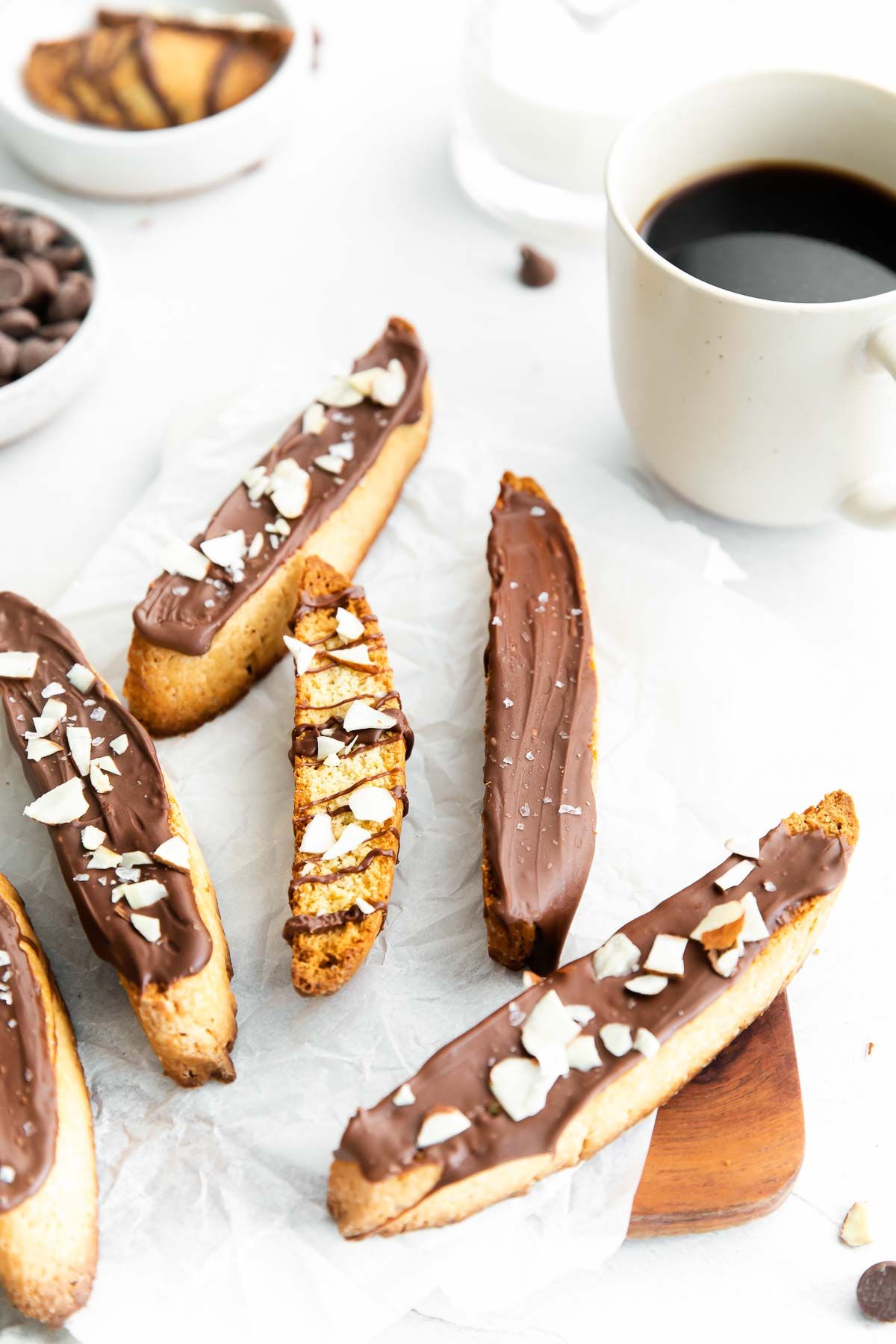 Vanilla biscotti covered in chocolate on a marble surface, bowl of chocolate chips and cup of coffee in the background.
