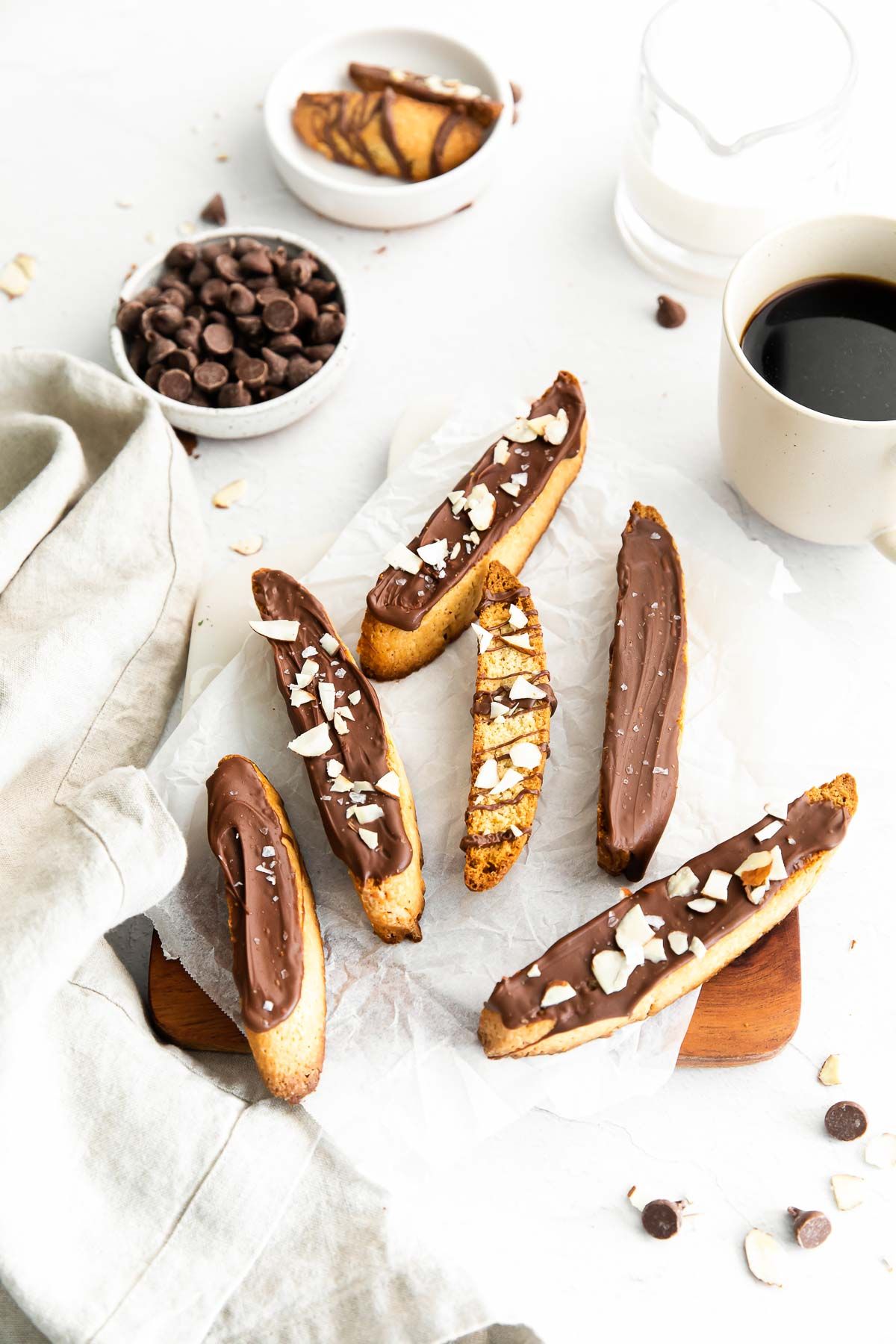 Vanilla biscotti covered in chocolate on a marble surface, bowl of chocolate chips and cup of coffee in the background.