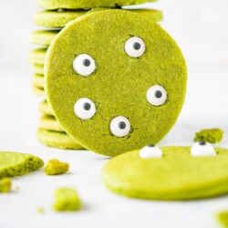 A round green Halloween Matcha cookie leaning against a stack of them against a white background. Edible eyes decorate the cookie to look like a green monster face.
