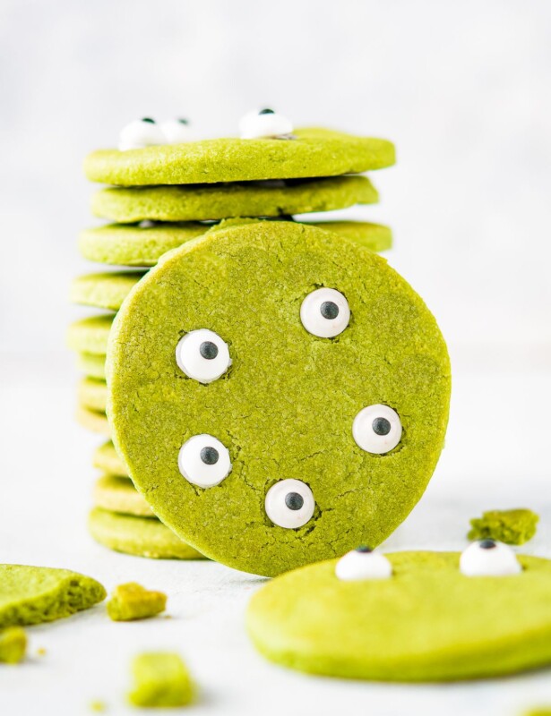 A round green Halloween Matcha cookie leaning against a stack of them against a white background. Edible eyes decorate the cookie to look like a green monster face.