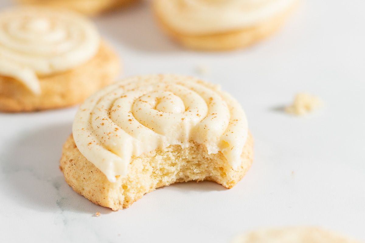 Cinnamon roll cookies swirled with cream cheese icing, on a white countertop. Bite removed from one.
