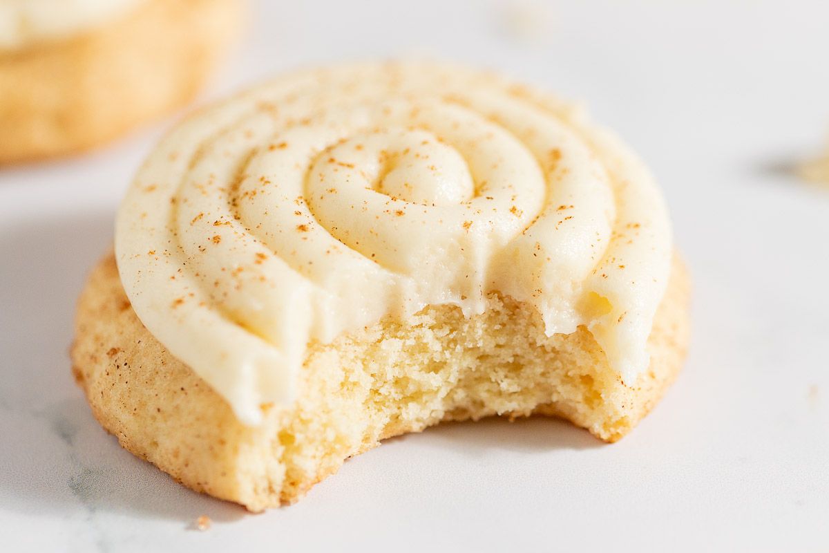 Cinnamon roll cookies swirled with cream cheese icing, on a white countertop. Bite removed from one.